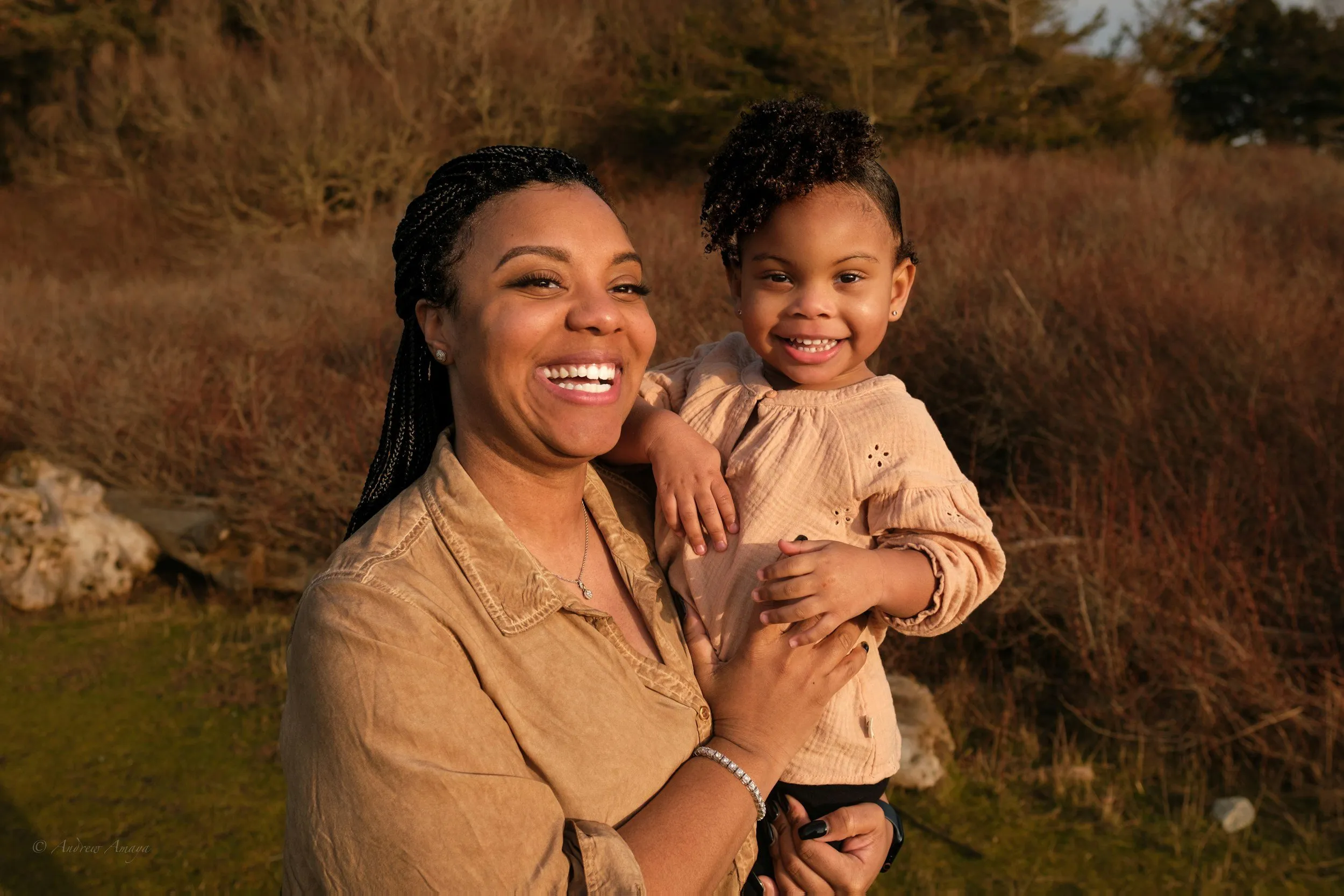 A smiling woman holding a toddler outdoors in golden light